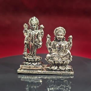 May include: Two silver-colored statues of Hindu deities. One statue is standing, the other is seated in a lotus position. Both statues are intricately detailed and rest on a shared base. The background is a gradient of red and black.
