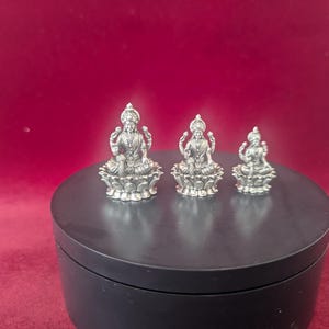 May include: Three silver figurines of the Hindu goddess Lakshmi sitting on lotus flower bases. The figurines are different sizes, with the largest one in the center.