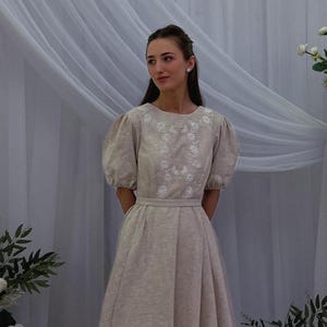 May include: A beige dress with short, puffed sleeves and a fitted waist. The bodice features white floral embroidery. The dress has a full skirt and is suitable for formal occasions. The dress is made of a soft, flowing fabric.