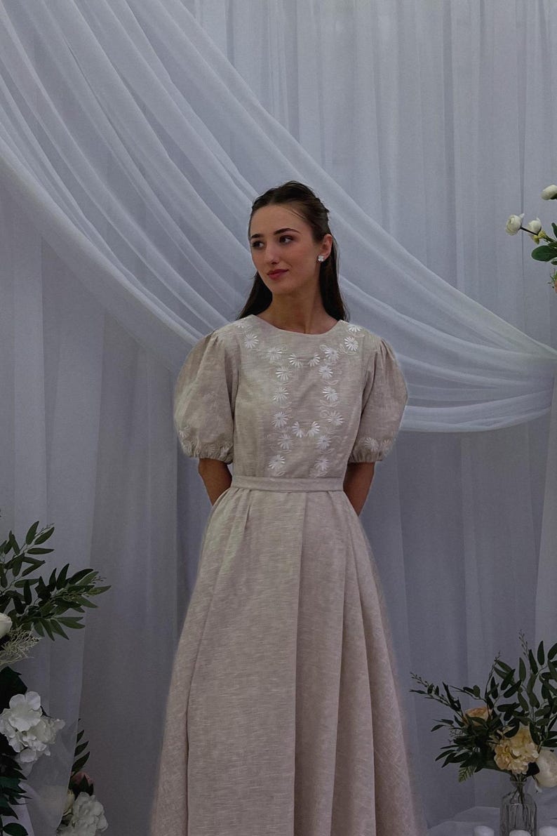 May include: A beige dress with short, puffed sleeves and a fitted waist. The bodice features white floral embroidery. The dress has a full skirt and is suitable for formal occasions. The dress is made of a soft, flowing fabric.