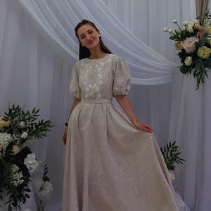 May include: A beige, floor-length dress with short, puffed sleeves and a fitted waist. The dress features embroidered floral detailing around the neckline. The woman is smiling and standing in front of a white curtain and floral arrangement.