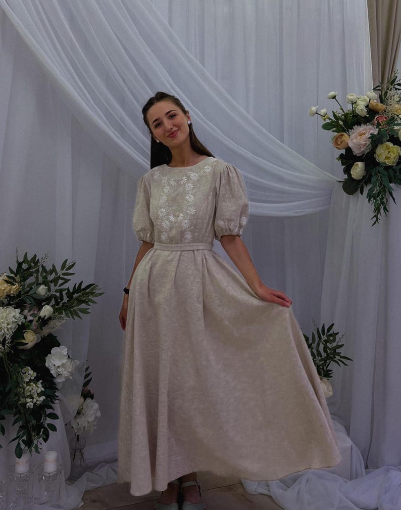 May include: A beige, floor-length dress with short, puffed sleeves and a fitted waist. The dress features embroidered floral detailing around the neckline. The woman is smiling and standing in front of a white curtain and floral arrangement.