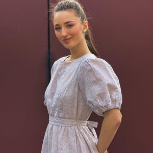 May include: A light brown dress with white floral embroidery on the bodice and sleeves. The dress features a square neckline, puffed sleeves, and a tie-waist detail. The dress is made of a textured fabric.