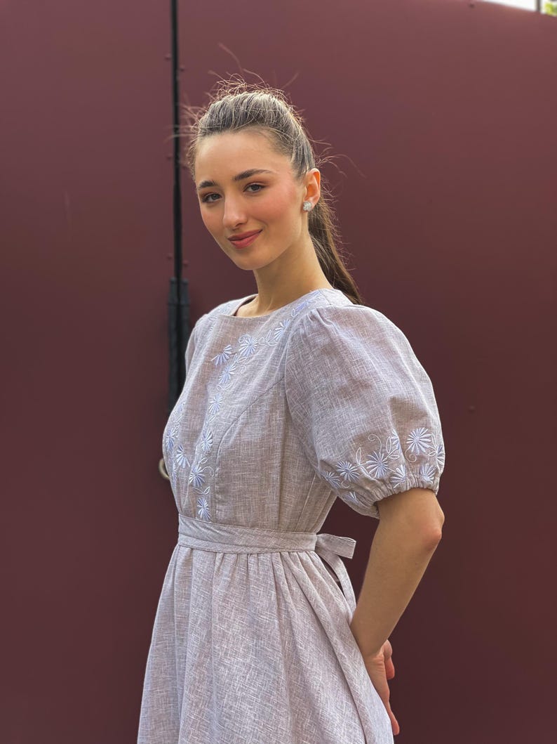 May include: A light brown dress with white floral embroidery on the bodice and sleeves. The dress features a square neckline, puffed sleeves, and a tie-waist detail. The dress is made of a textured fabric.