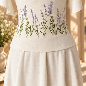 May include: A cream-colored dress featuring a fitted bodice with embroidered purple lavender sprigs and green stems. The garment has short flutter sleeves and a pleated skirt, crafted from a textured fabric with a visible weave pattern.