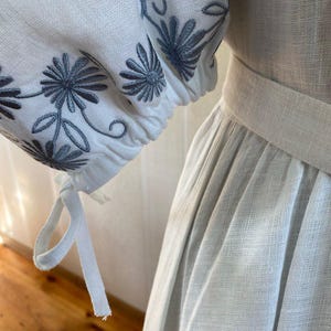 May include: Close-up of a white linen dress with embroidered sleeves. The sleeves feature a floral design in shades of gray. The dress has a fitted bodice and a flowing skirt, with a matching belt.