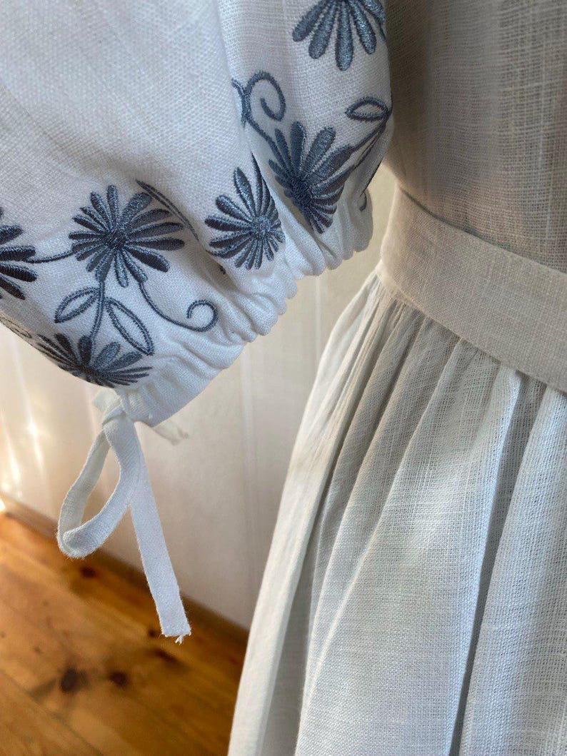May include: Close-up of a white linen dress with embroidered sleeves. The sleeves feature a floral design in shades of gray. The dress has a fitted bodice and a flowing skirt, with a matching belt.