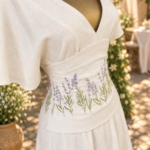 May include: A cream-colored dress featuring a deep V-neckline and wide flutter sleeves. The bodice is cinched with a wide waistband decorated with an embroidered pattern of purple lavender sprigs and green stems wrapping around the midsection.