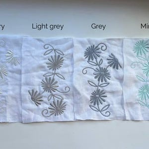 May include: A collection of white linen fabric swatches, each embroidered with a floral design in various colors. The colors include white, ivory, light grey, grey, mint, and blue. The text above each swatch indicates the color.