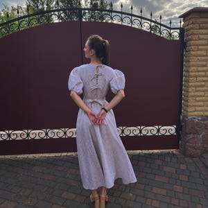 May include: A light-colored linen dress with short, puffed sleeves and a bow detail on the back. The dress has a fitted bodice and a flowing skirt. The person is wearing light-colored heeled sandals. The background includes a dark brown gate and a brick wall.