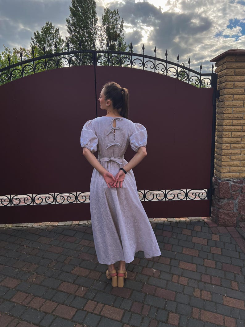 May include: A light-colored linen dress with short, puffed sleeves and a bow detail on the back. The dress has a fitted bodice and a flowing skirt. The person is wearing light-colored heeled sandals. The background includes a dark brown gate and a brick wall.