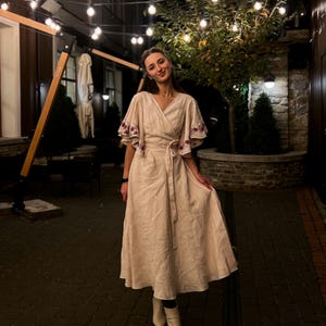 May include: A beige wrap dress with embroidered sleeves and a matching belt. The dress has a midi length and is paired with white ankle boots. The background features string lights and a stone wall, creating a warm, inviting atmosphere.