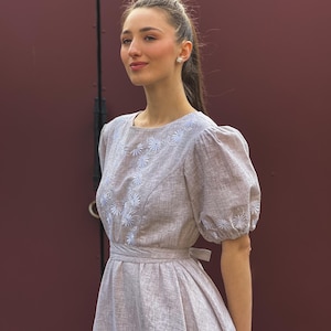 May include: A light brown dress with white floral embroidery. The dress features a round neckline, short puffed sleeves, and a belted waist. The dress is made of a textured fabric and has a knee-length skirt.