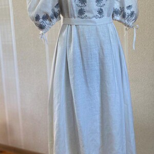 May include: A white linen dress with floral embroidery in a light blue hue. The dress features puffed sleeves, a cinched waist, and a flowing skirt. The embroidery is concentrated around the neckline and sleeves.