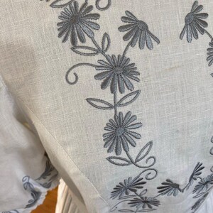 May include: Close-up of a white linen garment with gray floral embroidery. The embroidery features stylized flowers and leaves, creating a delicate pattern. The garment appears to be a dress or blouse, with visible seams and a soft texture.
