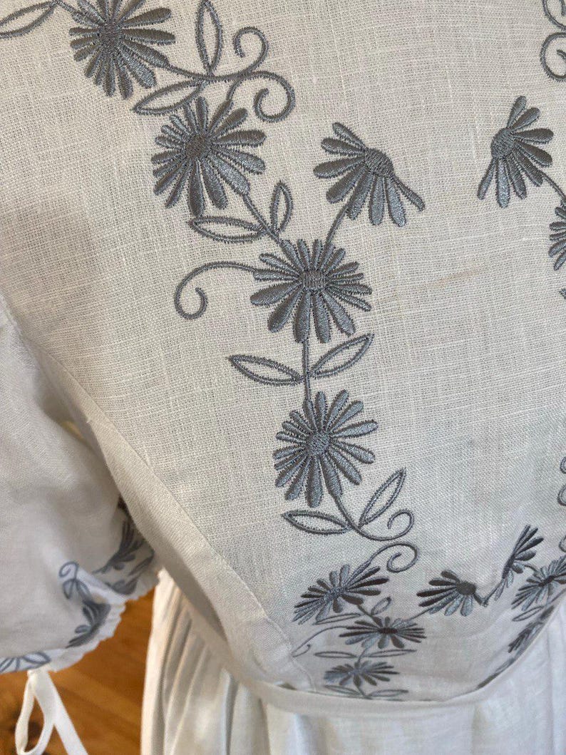 May include: Close-up of a white linen garment with gray floral embroidery. The embroidery features stylized flowers and leaves, creating a delicate pattern. The garment appears to be a dress or blouse, with visible seams and a soft texture.