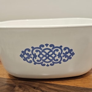 May include: A white oval casserole dish with integrated side handles, featuring a centered blue decorative filigree pattern on the front. The dish has a smooth, glossy finish and a deep basin, designed for oven use and serving food at the table.
