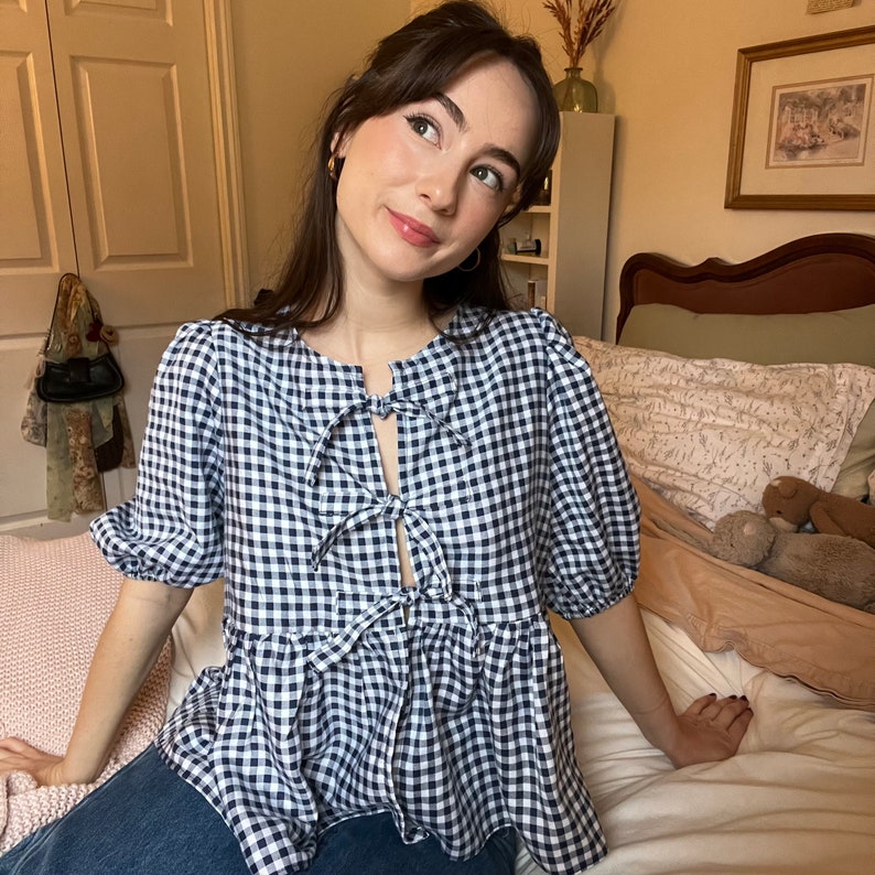 May include: A woman wearing a blue and white gingham top with a bow tie detail. The top has a ruffled hem and short sleeves. She is sitting on a bed with a white sheet and a brown pillow.