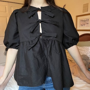 May include: A black, short-sleeved blouse with a gathered skirt and a unique bow detail at the neckline. The bow is made of multiple layers of fabric and is tied in a knot at the center.