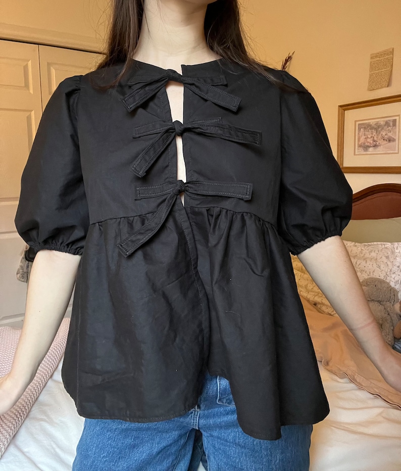 May include: A black, short-sleeved blouse with a gathered skirt and a unique bow detail at the neckline. The bow is made of multiple layers of fabric and is tied in a knot at the center.