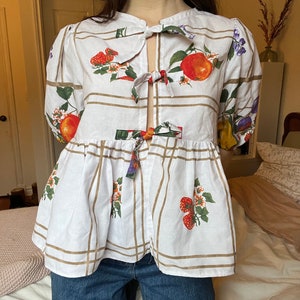 May include: A white blouse with a fruit print and a tie at the neckline. The blouse has a gathered skirt and brown stripes.