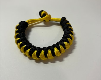 Può includere: Un braccialetto intrecciato nero e giallo realizzato con paracord.