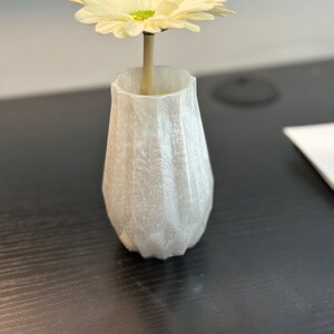 May include: A small, white, textured vase with a single, light yellow daisy. The vase has a geometric design and is sitting on a dark wood surface. The daisy has a green center.