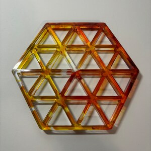 Puede incluir: Un salvamanteles hexagonal hecho de resina transparente con un degradado de amarillo, naranja y rojo. El salvamanteles está diseñado con un patrón geométrico de triángulos.