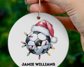 Op de afbeelding: Witte keramische kerstbal met een voetbal die een kerstmanmuts draagt en omringd is door kleurrijke kerstverlichting. De kerstbal is gepersonaliseerd met de naam "Jamie Williams" en het jaar "2024".