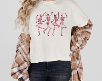 Puede incluir: Una camiseta blanca con un gráfico de cinco esqueletos con tutús rosas bailando. Los esqueletos están en varias poses, con los brazos y las piernas extendidos. La camiseta la lleva una persona con el pelo largo rubio y una camisa de cuadros.