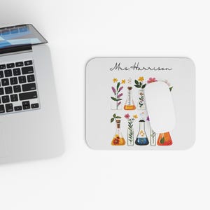 Pode incluir: Tapete de rato branco com um design floral com flores coloridas em frascos de laboratório. O tapete de rato tem o nome "Mrs. Harrison" em escrita cursiva. Um rato de computador branco repousa sobre o tapete, ao lado de um laptop.