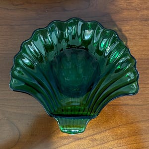 May include: A translucent emerald green glass dish shaped like a scallop shell. The dish features a series of radiating ridges that extend from the base to the scalloped edge, creating a textured, fan-like appearance with a smooth, concave center.