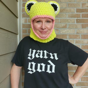 May include: A handmade, bright yellow crochet frog hat with black and white accents on the ears and a pink trim around the face opening. The person is wearing a black t-shirt with the words "yarn god" in white gothic lettering.