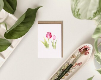 Könnte beinhalten: Eine Grußkarte mit einem Aquarellgemälde von zwei rosa Tulpen mit grünen Stielen und Blättern. Die Karte befindet sich in einem braunen Papierrahmen. Ein Pinsel liegt in einer dekorativen Blumenschale, mit grünem Laub im Hintergrund.