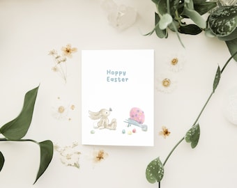 Może przedstawiać: Biała kartka z tekstem "Hoppy Easter" w niebieskich literach. Kreskówkowy królik trzyma niebieskiego motyla i patrzy na różowe jajko w taczce. Wokół królika i taczki rozrzucone są małe różowe i białe jajka.
