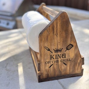 May include: A dark stained wooden paper towel holder with a handle. The side panel features a laser-engraved design of a crown, a grilling fork, and a spatula, with the text "KING OF THE GRILL" centered between the utensils.