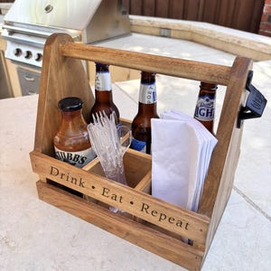 May include: A wooden condiment caddy with a handle and a black metal bottle opener on the side. The front is engraved with the text Drink • Eat • Repeat. It holds a bottle of Stubb's sauce, two beer bottles, clear plastic cutlery, and white napkins.