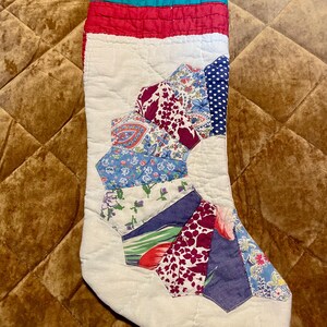 Può includere: Una calza natalizia trapuntata con base bianca e un design patchwork a ventaglio composto da vari segmenti di tessuto floreale e fantasia in tonalità blu, rosso e viola. I bordi superiore e inferiore presentano fasce color ottanio e rosso.