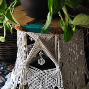 May include: A handcrafted macrame side table with a wooden top. The table features a natural wood slice top with a teal resin inlay. The macrame base is made of cream-colored rope and has a geometric design with hanging crystal accents.