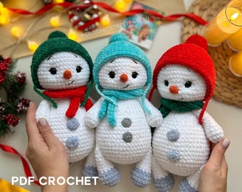 May include: Three handmade crochet snowmen with white bodies, each wearing a different colored hat and scarf: green, blue, and red. The snowmen have black eyes, orange noses, and gray button accents. The text "PDF CROCHET PATTERN" is visible.