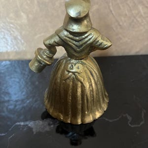 Op de afbeelding: Een vintage messing beeldje van een vrouw in een lange jurk en hoed, die een klein voorwerp vasthoudt. De figuur heeft ingewikkelde details en een warme, gouden kleur. Het beeldje is waarschijnlijk een decoratief item.