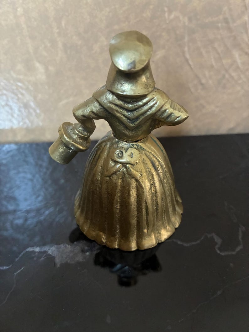 Op de afbeelding: Een vintage messing beeldje van een vrouw in een lange jurk en hoed, die een klein voorwerp vasthoudt. De figuur heeft ingewikkelde details en een warme, gouden kleur. Het beeldje is waarschijnlijk een decoratief item.