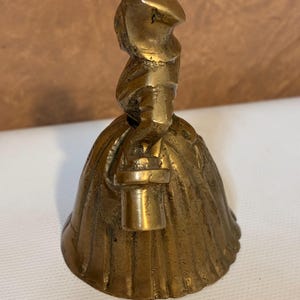 Op de afbeelding: Een vintage messing bel in de vorm van een vrouw in een lange jurk en hoed, met een lantaarn. De bel heeft een gedetailleerd ontwerp en een warme, gouden kleur. De bel is waarschijnlijk een verzamelobject.