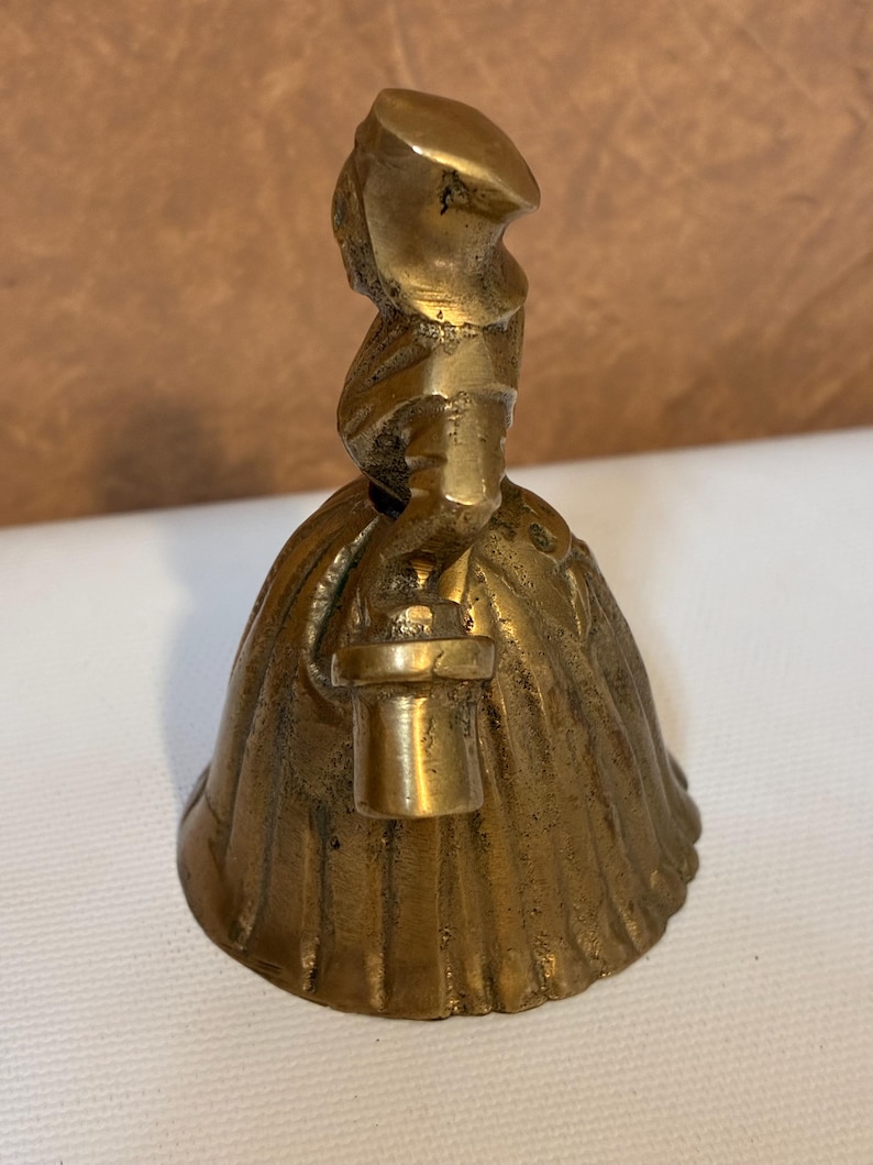 Op de afbeelding: Een vintage messing bel in de vorm van een vrouw in een lange jurk en hoed, met een lantaarn. De bel heeft een gedetailleerd ontwerp en een warme, gouden kleur. De bel is waarschijnlijk een verzamelobject.