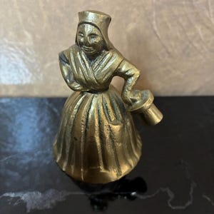 Op de afbeelding: Een kleine messing figuur van een vrouw in een traditionele muts en jurk, die een klein voorwerp vasthoudt. Het beeldje heeft een gedetailleerd ontwerp en een gepolijste afwerking. De figuur is ongeveer 7.6 cm hoog.