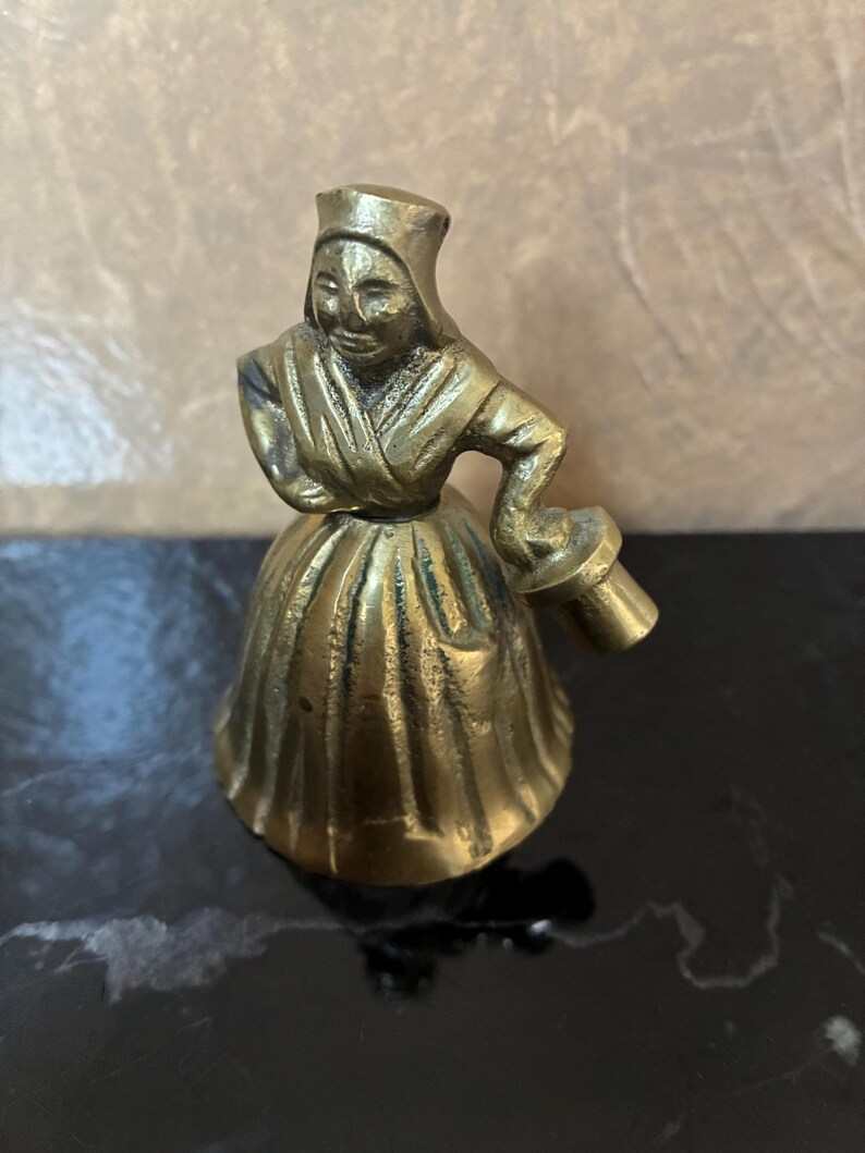 Op de afbeelding: Een kleine messing figuur van een vrouw in een traditionele muts en jurk, die een klein voorwerp vasthoudt. Het beeldje heeft een gedetailleerd ontwerp en een gepolijste afwerking. De figuur is ongeveer 7.6 cm hoog.