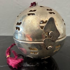 May include: A silver-colored, spherical decorative object with cut-out designs. It has a hinged closure and a small, maroon tassel. The object is likely made of metal and has a vintage or antique appearance.