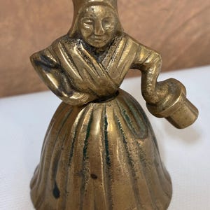 Op de afbeelding: Een vintage messing bel in de vorm van een vrouw in traditionele kleding en hoed. De bel heeft een handvat in de vorm van een kleine emmer. Het messing heeft een warme, gouden kleur met wat groene oxidatie.