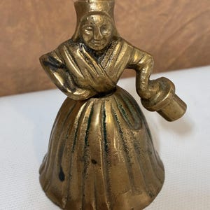 Op de afbeelding: Een vintage messing bel in de vorm van een vrouw in een traditionele jurk. De bel heeft een gedetailleerd gezicht en houdt een klein voorwerp vast. Het messing heeft een warme, verouderde patina en de bel staat op een wit oppervlak.