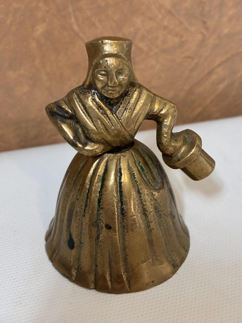 Op de afbeelding: Een vintage messing bel in de vorm van een vrouw in een traditionele jurk. De bel heeft een gedetailleerd gezicht en houdt een klein voorwerp vast. Het messing heeft een warme, verouderde patina en de bel staat op een wit oppervlak.